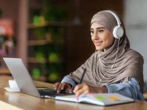 A woman in hijab wearing headphones and smiling at the screen